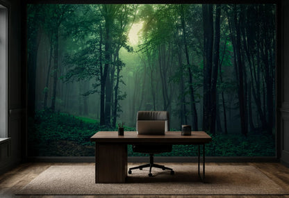 A dark wood desk with a laptop and two small cups placed in front of a large wall mural depicting a misty green forest with sunlight filtering through the trees.