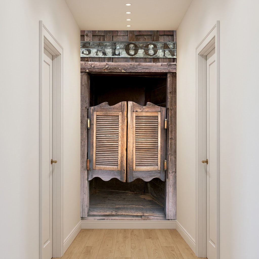 A narrow hallway with white walls and light wood flooring, featuring a large rustic wooden door or doorway centered in the frame. The door has louvered panels and a weathered wood finish, with a dark sign above reading 'SALOON'.