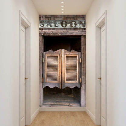 A narrow hallway with white walls and light wood flooring, featuring a large rustic wooden door or doorway centered in the frame. The door has louvered panels and a weathered wood finish, with a dark sign above reading 'SALOON'.