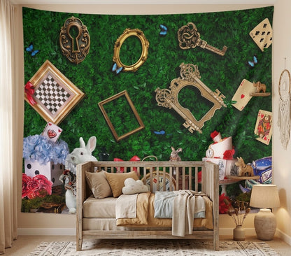 A nursery room wall decorated with a large green fantasy-themed tapestry featuring vintage gold frames, keys, playing cards, a chessboard, a white bunny, and whimsical illustrations. In front of the tapestry is a wooden crib with beige and tan bedding, flanked by a side table with a lamp and other nursery decor.