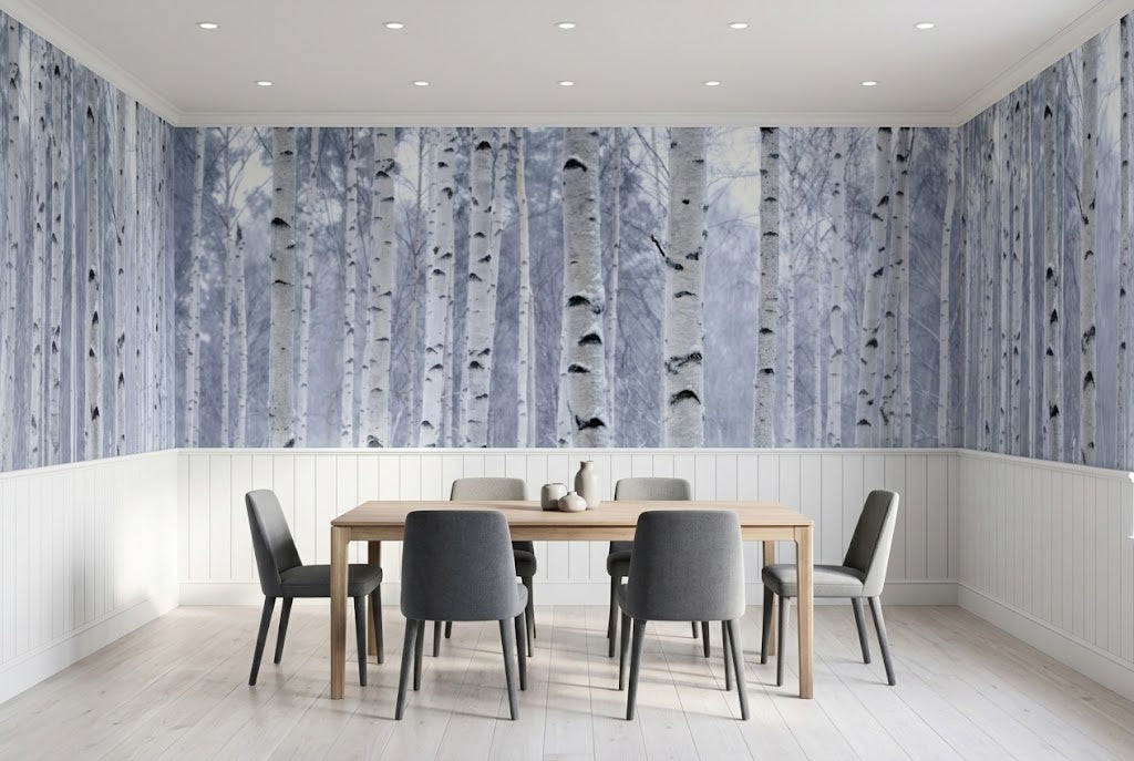 A dining room with a large wall mural depicting vertical birch tree trunks in shades of blue, white, and gray. The room features a light wood dining table and six gray upholstered chairs, set against a white wainscoted wall and light wood flooring.