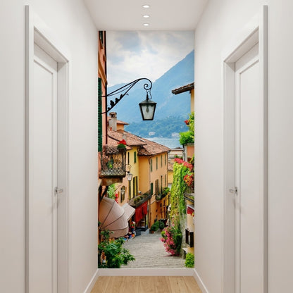 A hallway with a large wall mural of a scenic Italian village street with colorful buildings, a black street lamp, and mountains in the background.