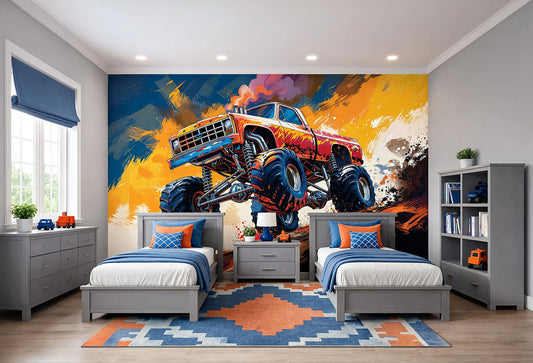 A children's bedroom featuring a large wall mural of a red and blue monster truck racing on a yellow and orange paint-splattered background. The room is styled with two gray single beds, blue and orange pillows, a geometric blue and orange rug, a gray dresser, and a gray bookshelf.