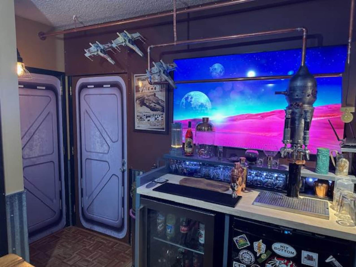 A themed bar setup with a large backlit wall display showing a colorful outer-space landscape, two sci-fi style doors on the left, a rocket-shaped tap, and various bar accessories.
